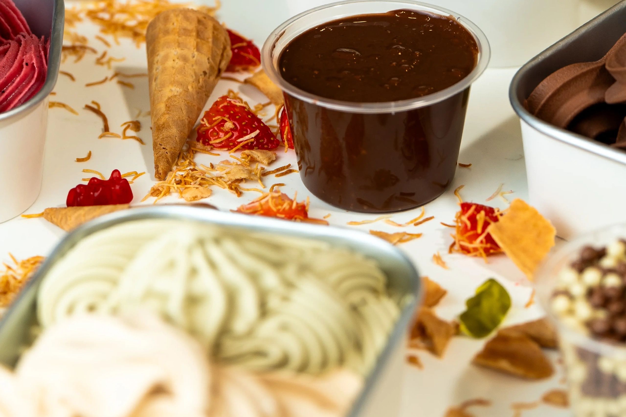 Small cup of chocolate sauce surrounded by ice cream cones and toppings.