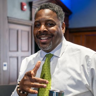 Smiling man in white shirt and green tie pointing at the camera indoors.