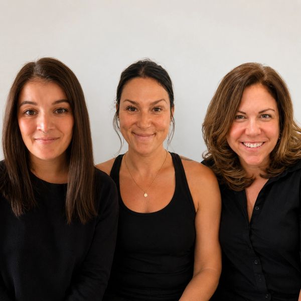 Three women smiling, dressed in black, against a plain light background.