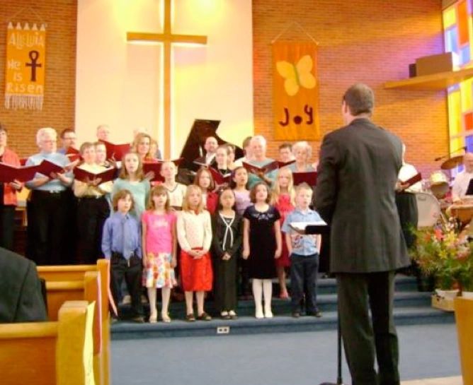 Children and adults singing in a church choir led by a conductor.