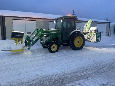 Green tractor with snow-clearing attachments on a snowy driveway.