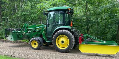 John Deere with a land plane, loader and grapple bucket, on freshly graded gravel driveway.