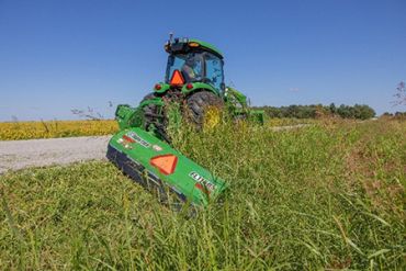 Green tractor mowing tall grass beside a gravel road under a clear blue sky.