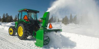 Green tractor clearing snow with a snow blower on a snowy road.