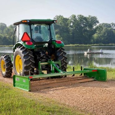 A tractor grading a gravel path near a lake with fishermen in a boat.