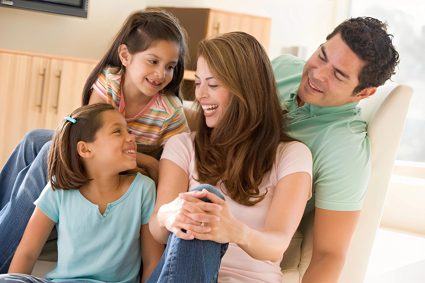 A smiling family of four, including two parents and two young daughters, sits together on a couch.