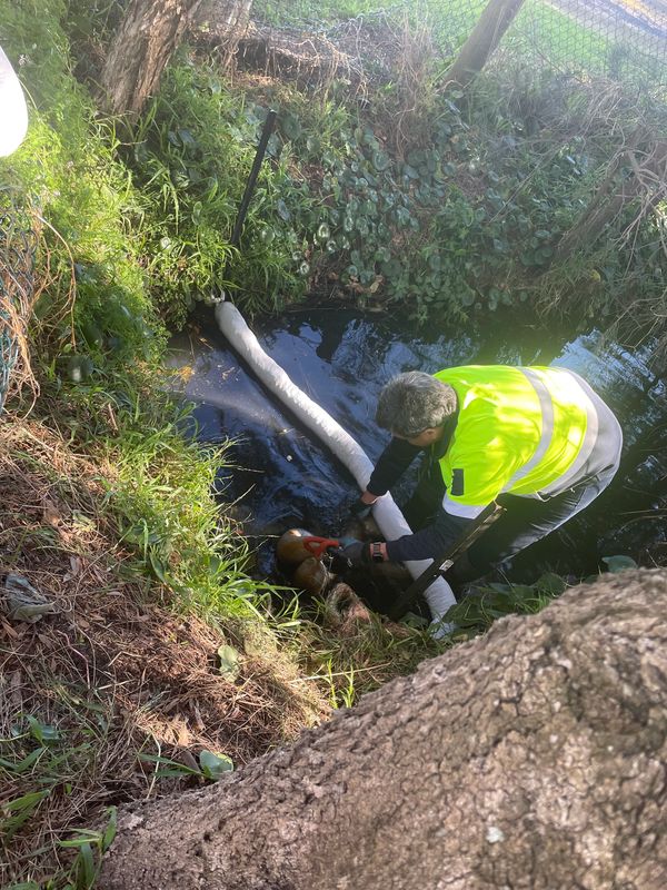 Worker in high-visibility gear cleans a small waterway with equipment.