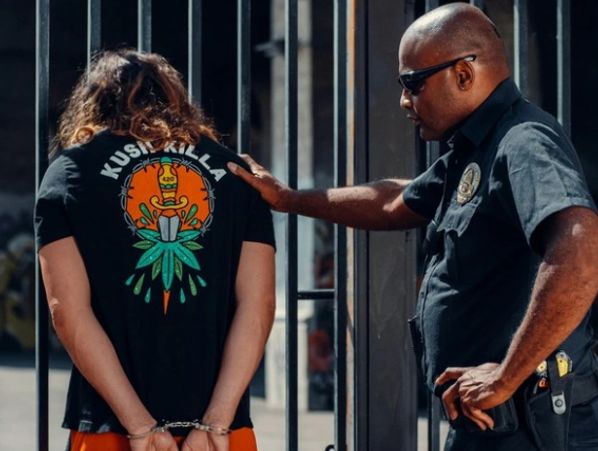 Police officer detains a handcuffed man wearing a "Kush Killa" t-shirt.