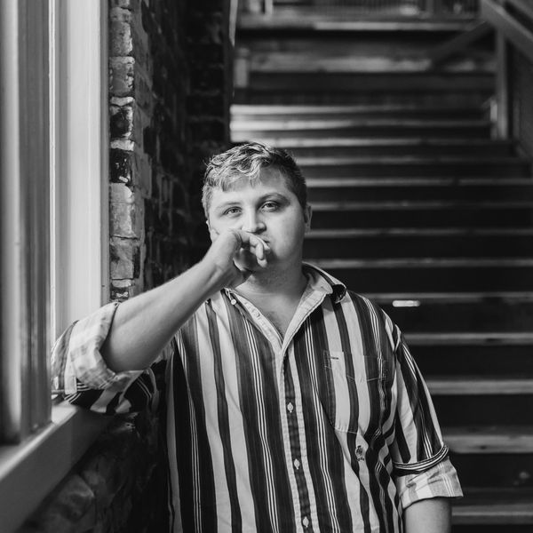 Man in a striped shirt leaning by a window, looking thoughtful.