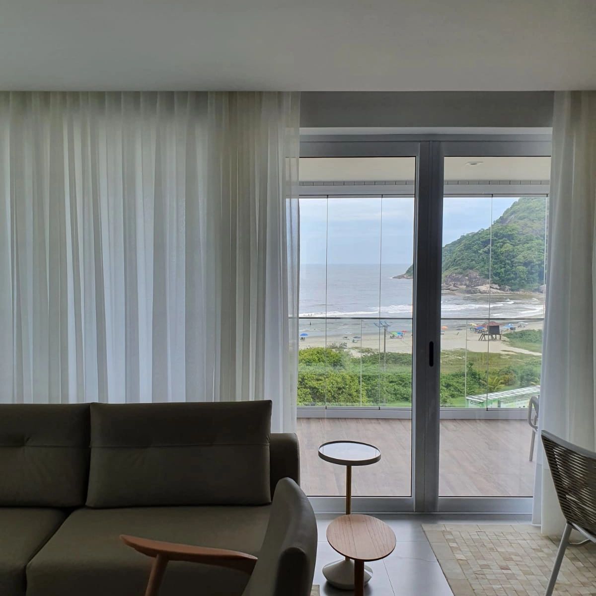 Ambiente com vista para o mar, com cortinas brancas, sofá e poltrona cinza. Cortina wave em gaze.