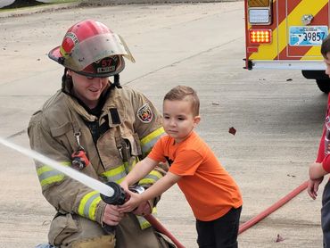Firefighter helps young boy spray water from a hose.