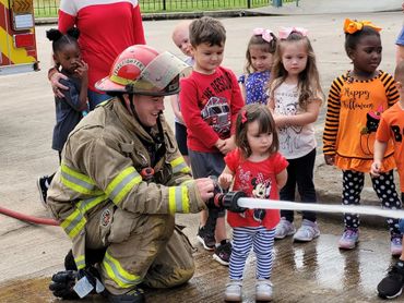 Firefighter helps young girl spray water from a fire hose while children watch.