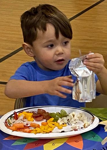 Young boy in blue shirt opening a juice pouch at a colorful table with a plate of snacks.