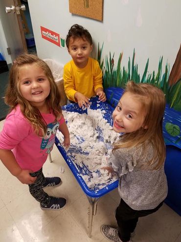 Three children playing happily with a sensory table filled with white material.