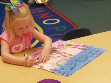 Young girl coloring a colorful drawing at a classroom table.