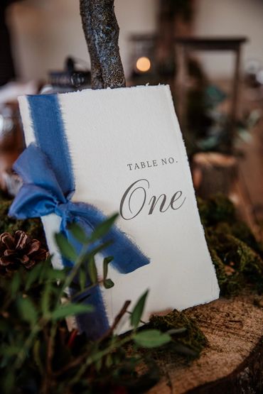 White table number sign with a blue ribbon, leaning against a log