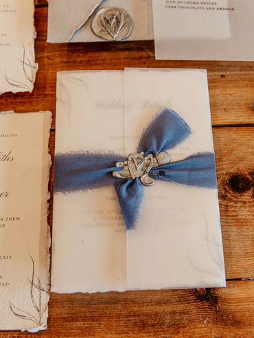 Wedding Invitation with a vellum wrap, blue ribbon and a grey wax seal, lay on wooden table