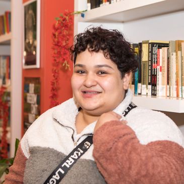 a mid-30s person, with short hair and a cozy V neck zip sweater, standing in front of a bookshelf