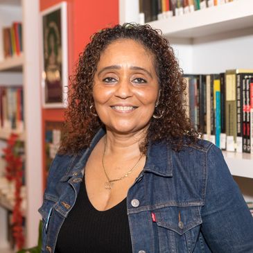 middle aged woman with curly hair and a jean jacket, standing in front a book shelf