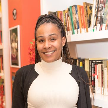 a young 30 or 40s aged black woman wearing a cardigan and standing in front of book shelf