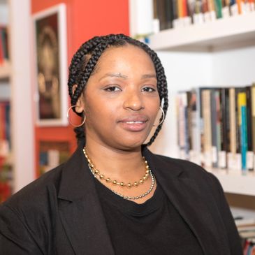 black woman in her mid 30s, with braids held back, golden jewelry, a slight smile