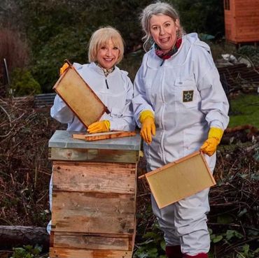 Esther and Jane Horrocks are wearing bee suits, standing behind a beehive and holding frames