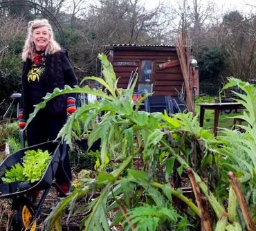 Esther holds a wheelbarrow in her allotment with a shed behind her and plants in front of her