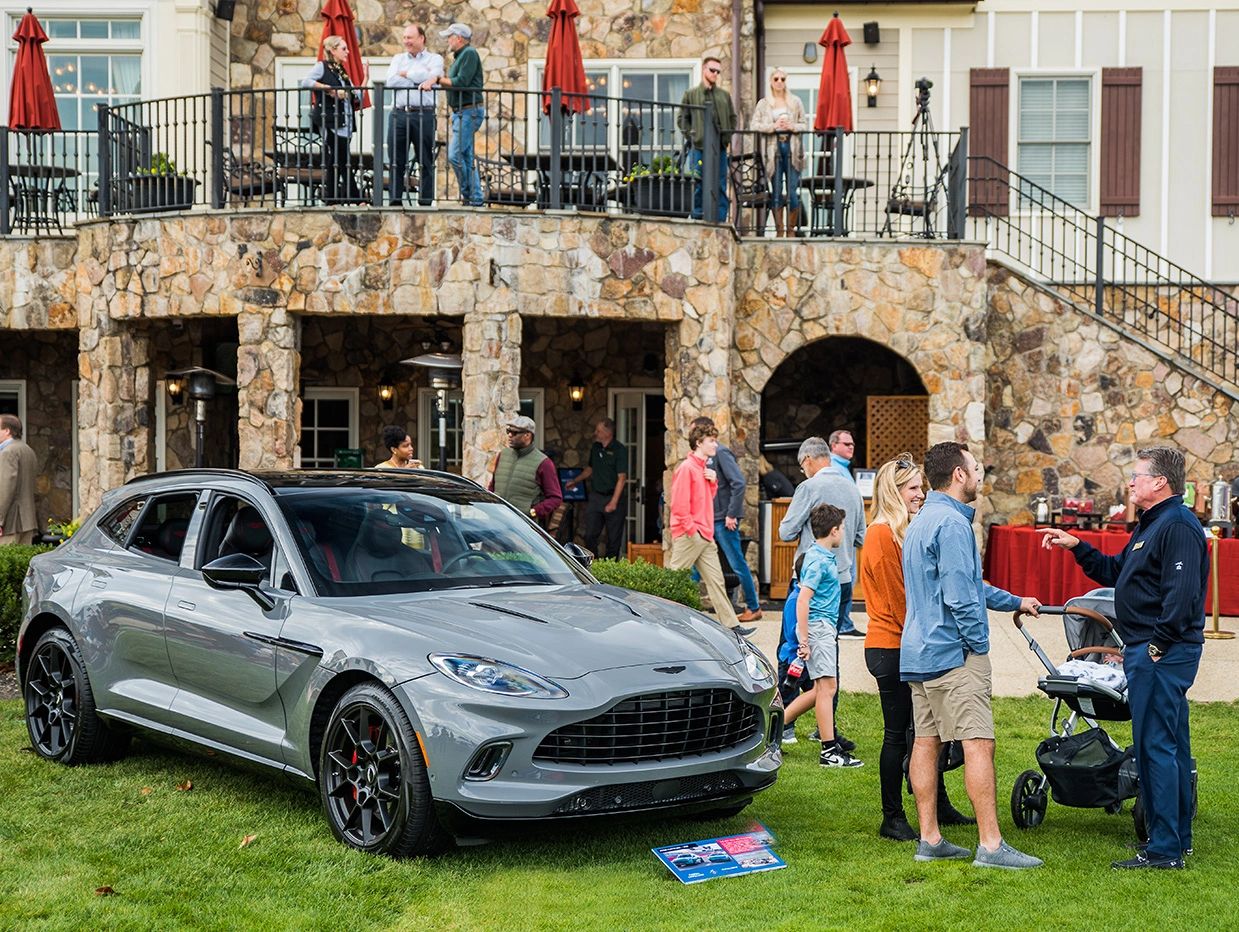 A silver luxury car displayed outdoors with people conversing nearby.