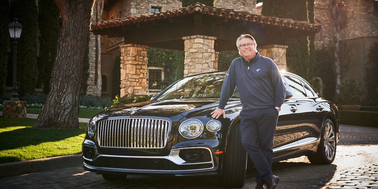 A man leaning on a luxury black car outside a stone building.