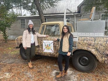 Two women standing beside a camouflaged truck with a hunting plantation logo.