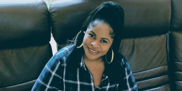 A smiling woman in a blue plaid shirt sitting on a brown leather couch, black leather sofa.