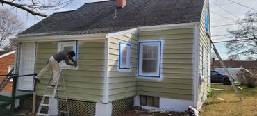 Man painting the exterior of a house, preparing windows with tape and plastic.