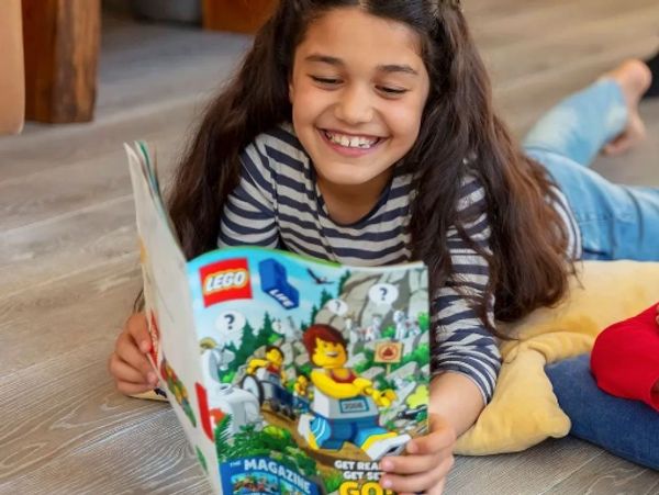 A girl happily reads a colorful LEGO magazine while lying on the floor.