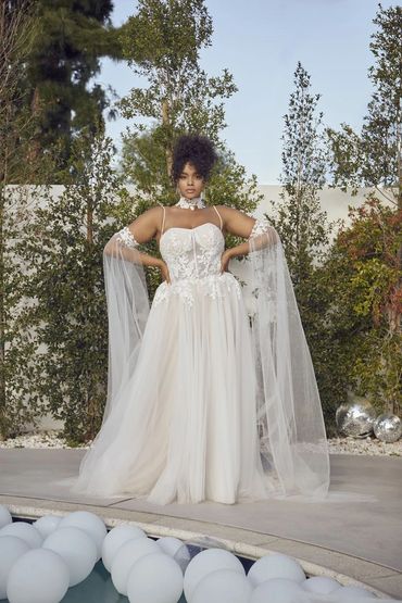 Woman in elegant white lace gown stands outdoors by pool and balloons.
