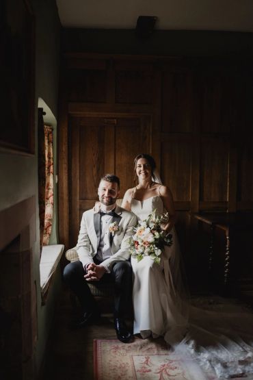 Newlywed couple posing indoors with bouquet and elegant attire.
