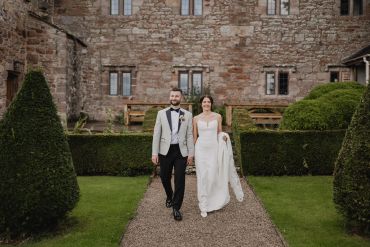 Newlywed couple walking in a garden with stone building backdrop.