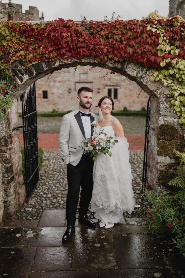 Bride and groom posing under a stone arch with autumn leaves.