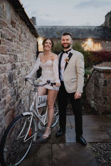Bride and groom smiling by a bicycle with wedding sign.