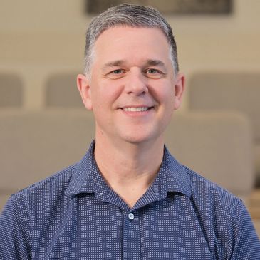 Smiling middle-aged man in a blue patterned shirt in a softly lit room.