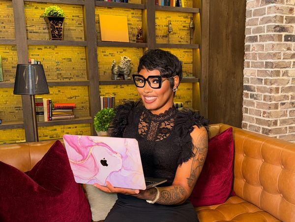 Woman in black dress with laptop sitting on a brown leather couch in a cozy library.