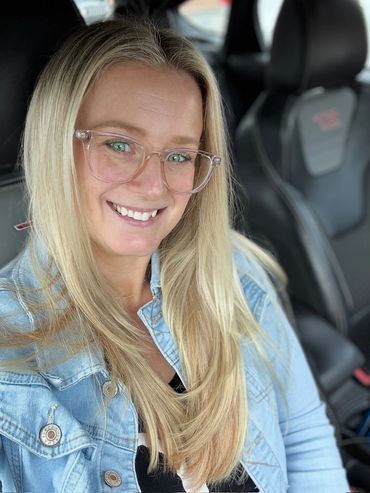Smiling woman with glasses wearing a denim jacket inside a car.