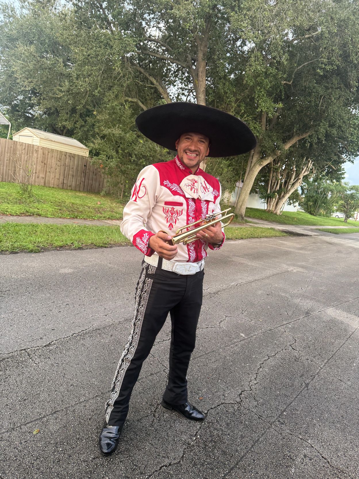 Man in traditional mariachi outfit holding a trumpet, smiling outdoors.