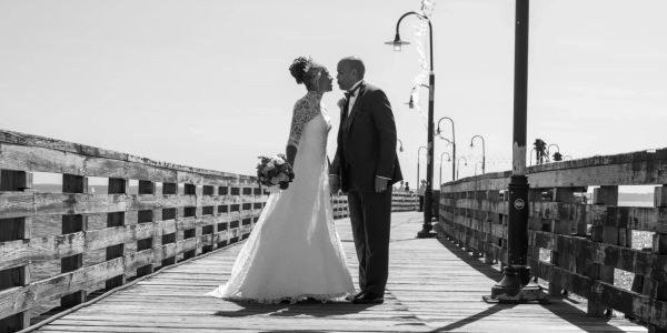Black and white photo of a bride and groom about to kiss on a wooden pier.