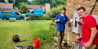 Two electricians working on an electrical panel outside a brick house.