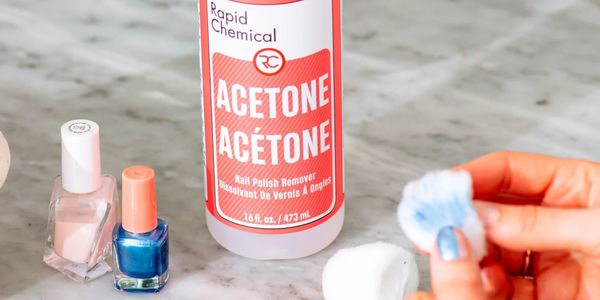 Bottle of acetone nail polish remover with cotton balls and nail polish bottles on a marble surface.