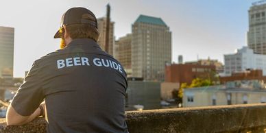 A man wearing a 'Beer Guide' shirt overlooking a cityscape at sunset.