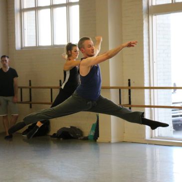 Two dancers performing a split leap in a studio.