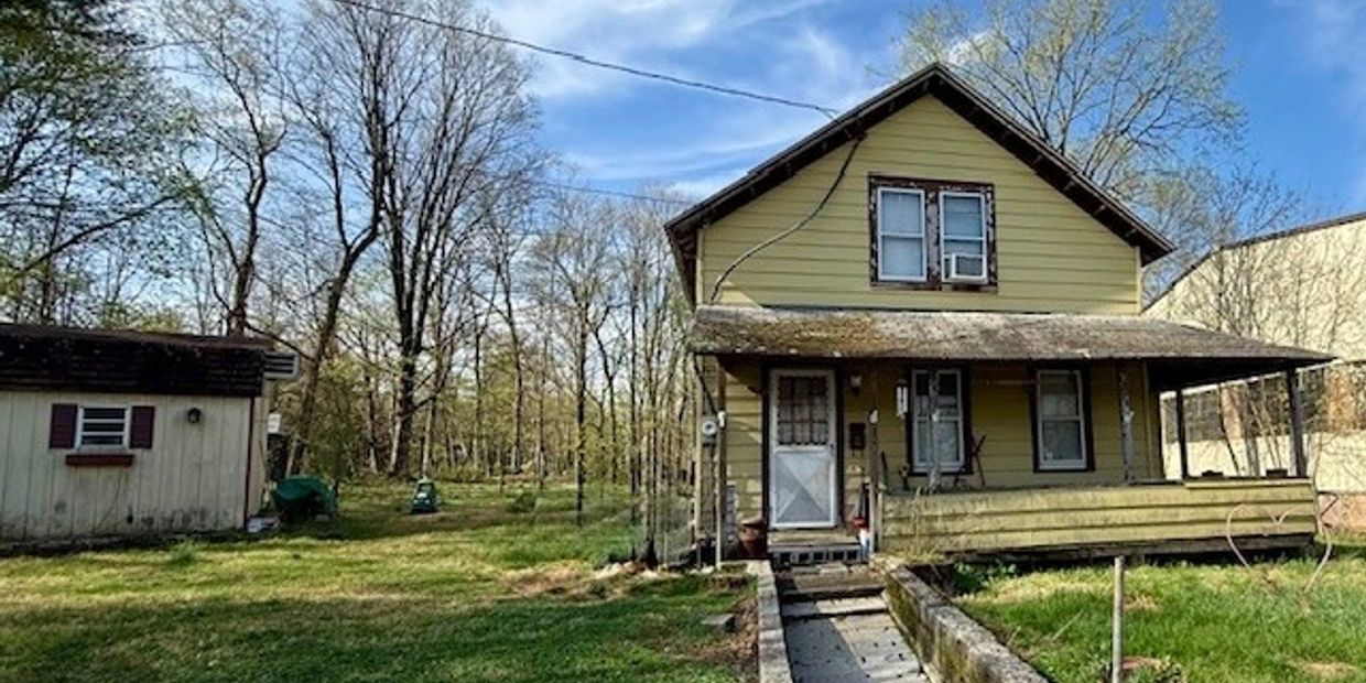 Old yellow house with a porch and a small shed in the yard on a sunny day.