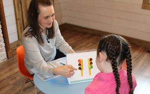 A speech language pathologist sits at a colorful table with while performing an assessment. 