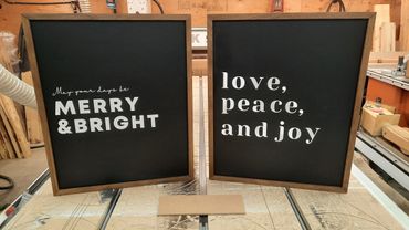 Two framed signs with festive holiday messages on a workshop table.